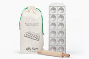 Metal ravioli mold with 12 hemispherical cavities, wooden rolling pin, and canvas storage pouch with green drawstring.