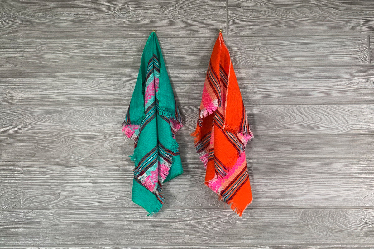 Two folded napkins in green and orange with geometric patterns and pink fringe trim hanging against gray wood background.