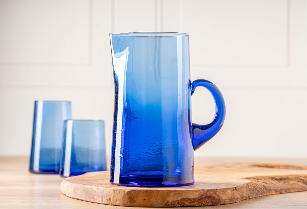 Three blue glass jugs with cone-shaped bodies, including one large jug with handle and two smaller glasses, on wooden coaster