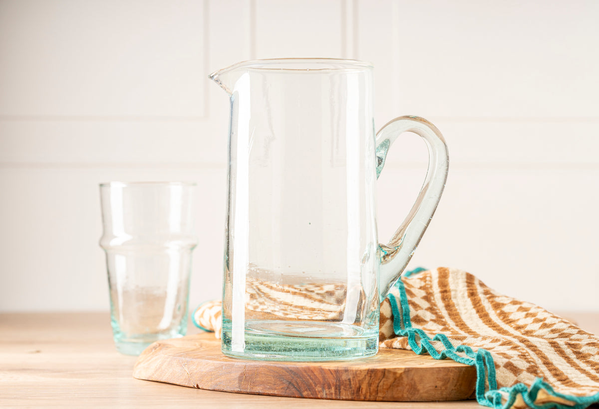 Clear glass pitcher with handle and spout beside matching glass, on wooden board with striped textile.