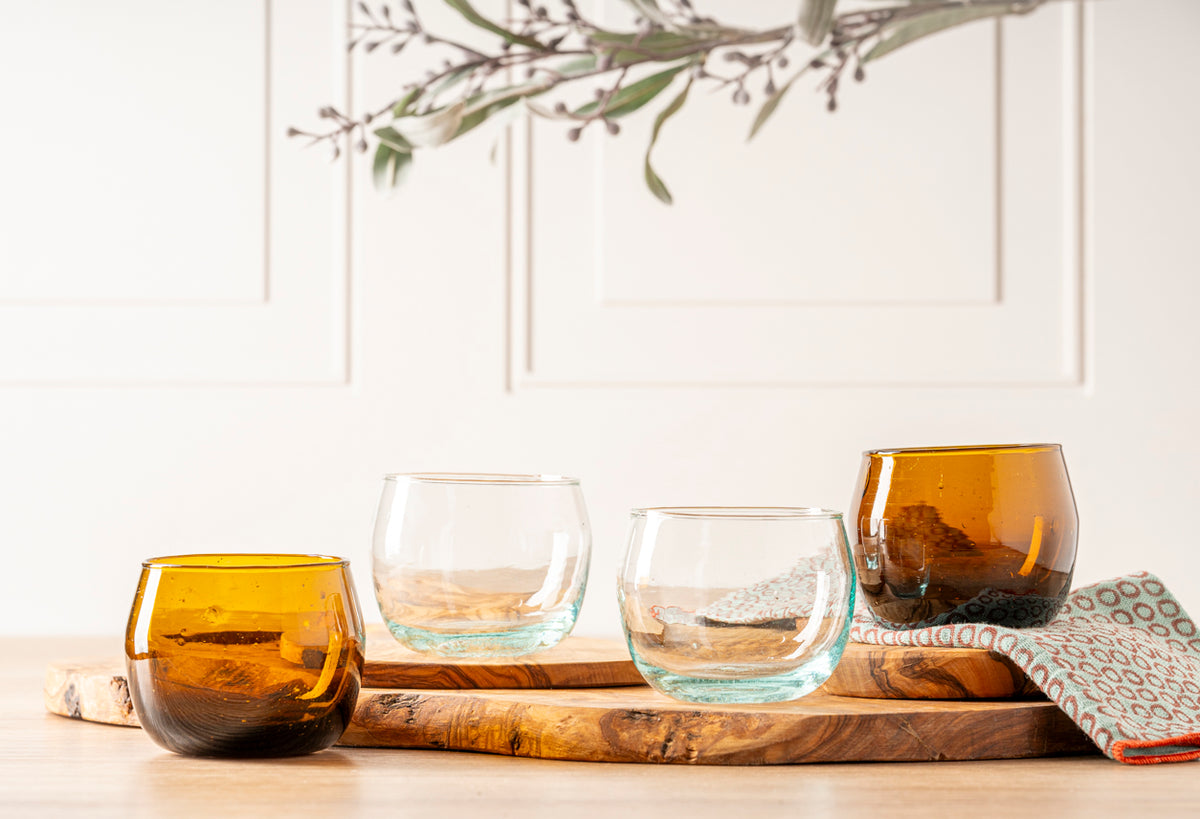 Four stemless goblets with amber and clear glass displayed on wooden tray with decorative cloth.