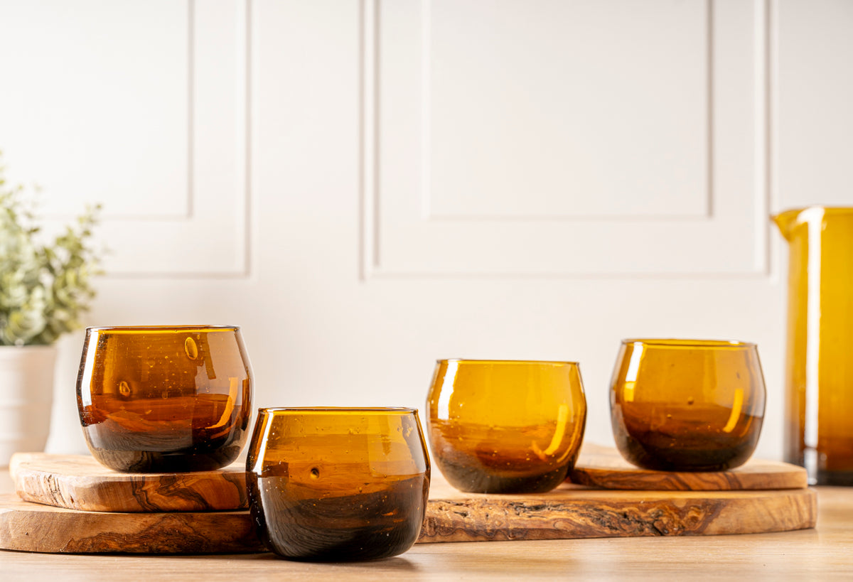 Four amber-colored stemless goblets with curved sides displayed on wooden surfaces.