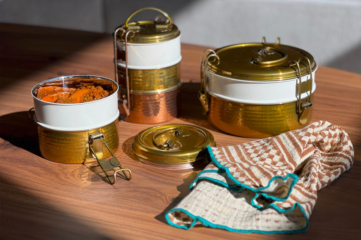 Three-tier stainless steel tiffin lunch box with brass handles and lids, one container filled with curry, displayed with clot