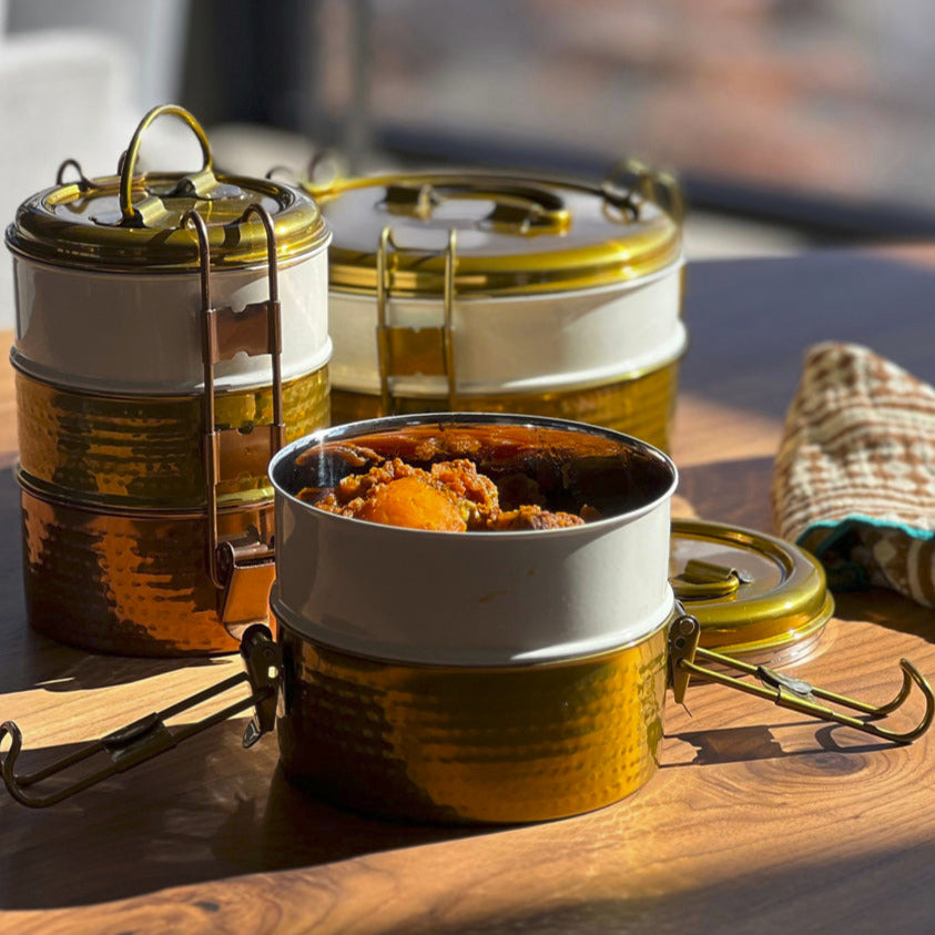 Stainless steel tiffin lunch box with three stacked compartments, brass handles, and lids containing food.