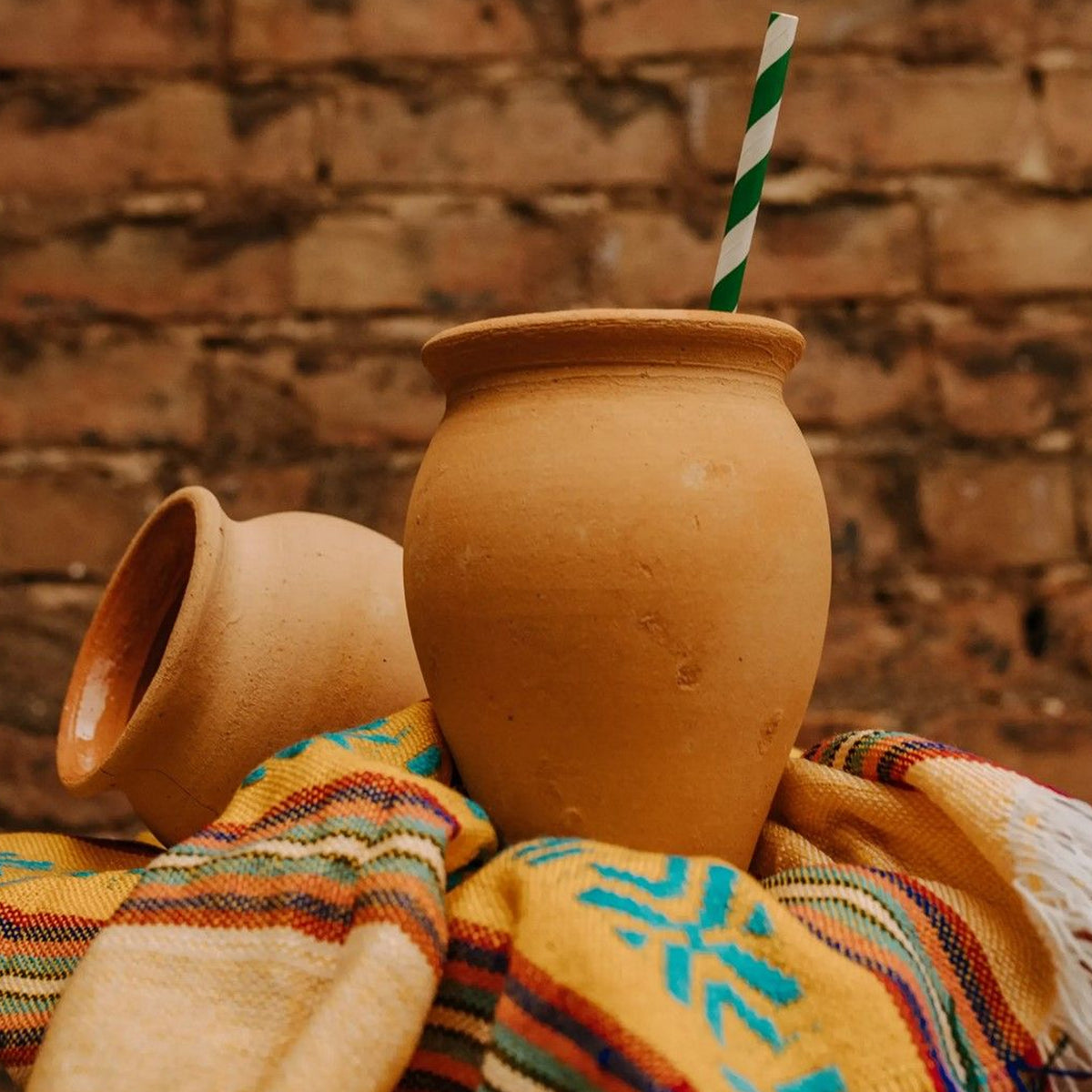 Two terracotta clay cups with wooden rim and green striped straw, displayed on colorful woven fabric against a brick wall.