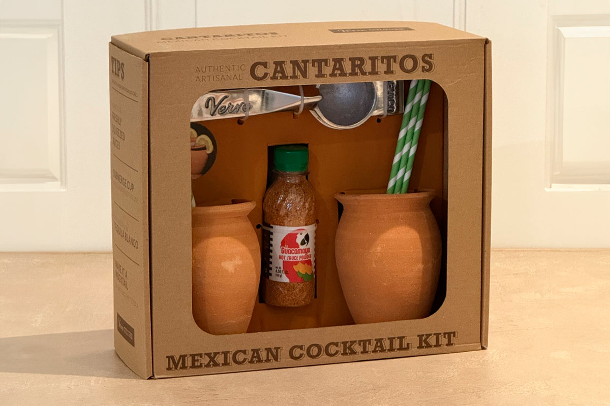 Cantaritos kit in cardboard box containing two terracotta cups, bottle of seasoning mix, striped straws, and bar spoon.