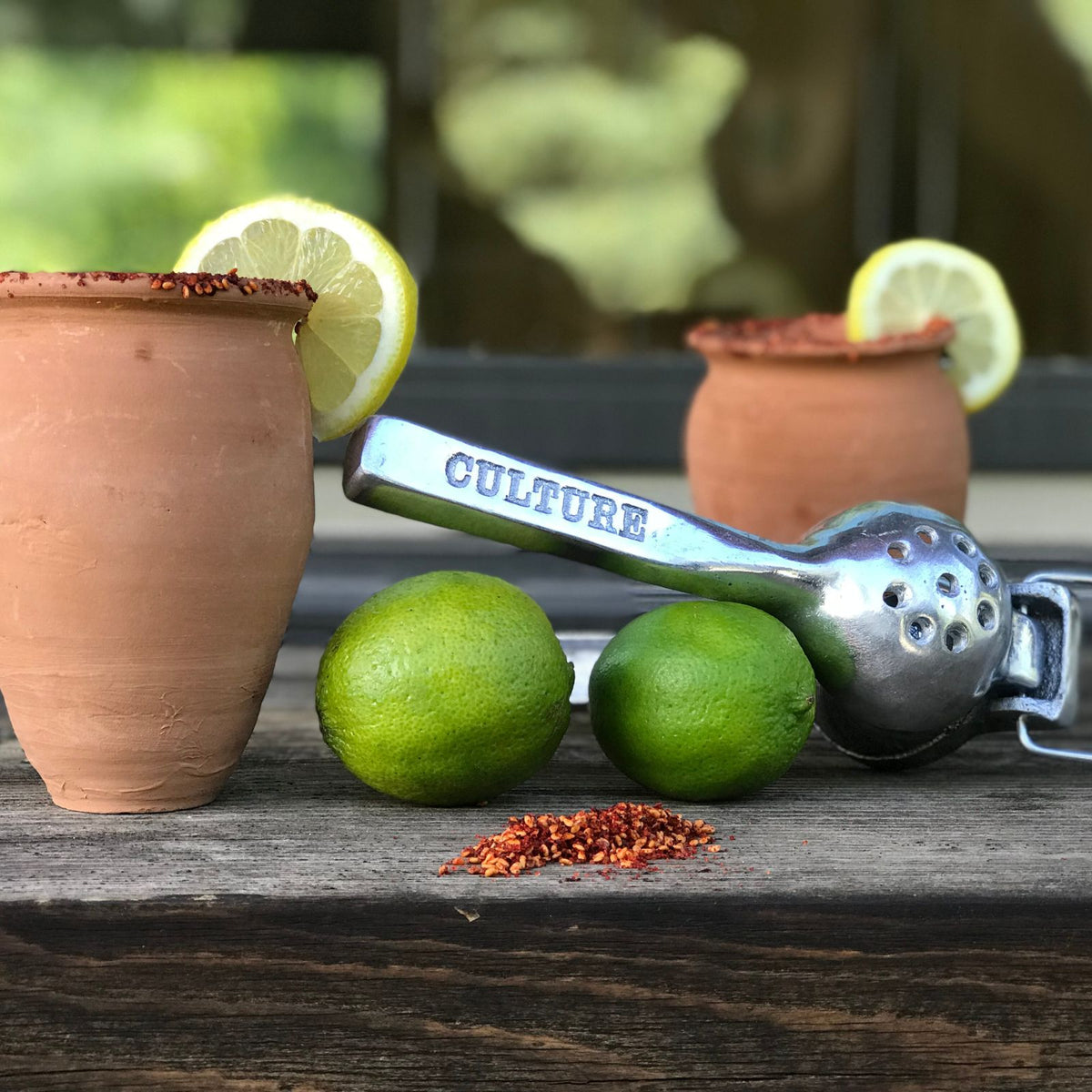Two terracotta cups with lime slices and chili rim, accompanied by limes, a metal juicer, and spoon on wooden surface.
