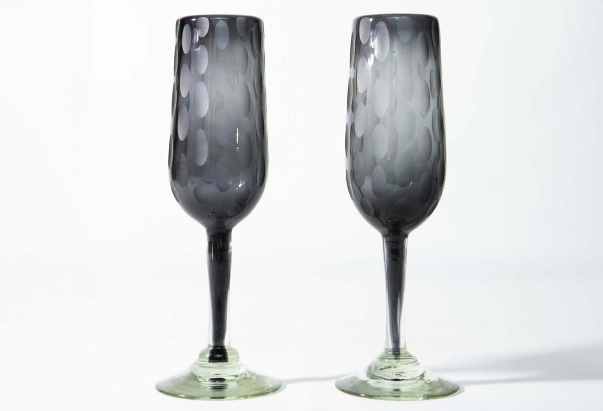 Two dark gray handblown champagne flutes with etched oval patterns and green glass bases.