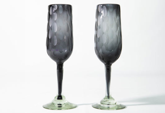 Two dark gray handblown champagne flutes with etched oval patterns and green glass bases.