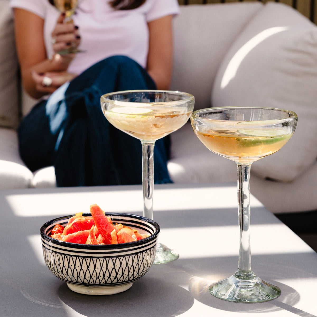 Two clear handblown cocktail glasses with long stems, each filled with light-colored beverage and garnished with citrus.