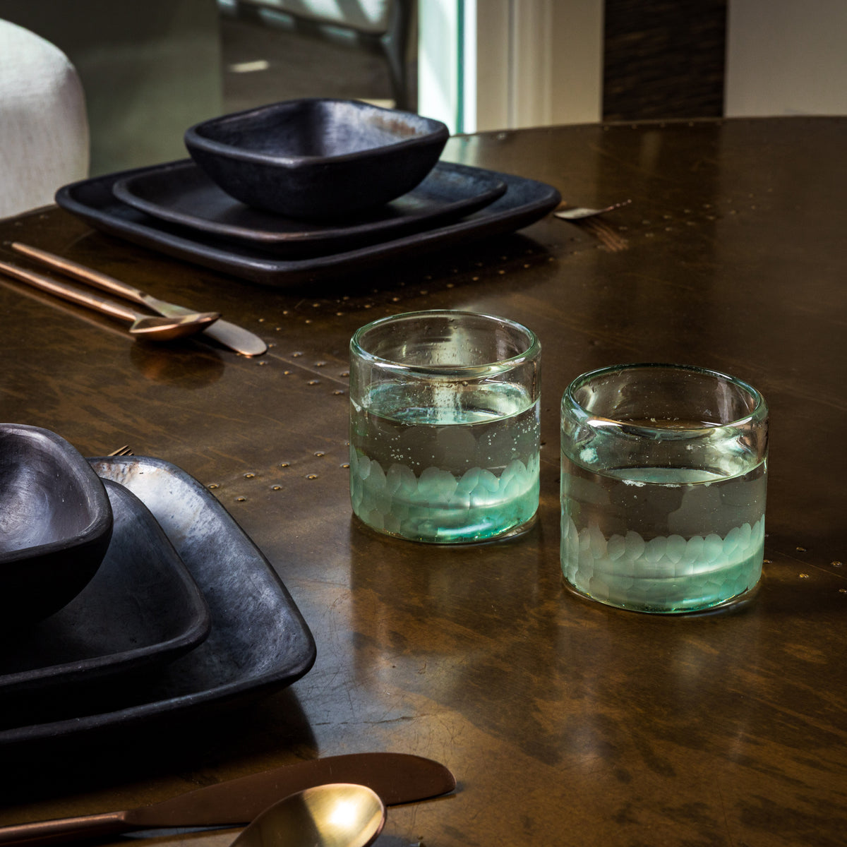 Two clear rocks glasses with etched green and white patterns, filled with ice water, on a dark table.