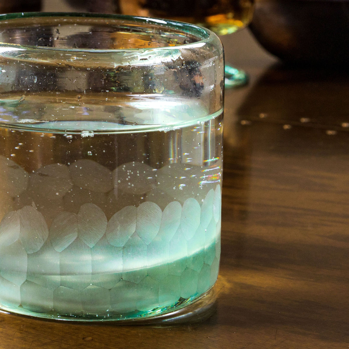 Handblown rocks glass with etched leaf pattern band, filled with clear liquid, shown on wooden surface.