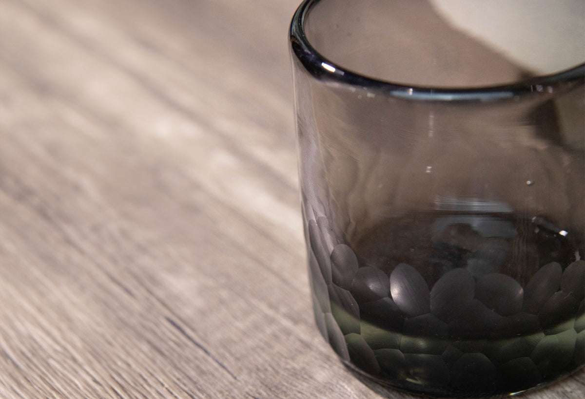 Smoky gray handblown glass rocks glass with etched texture on lower half and dark rim, shown on wooden surface.