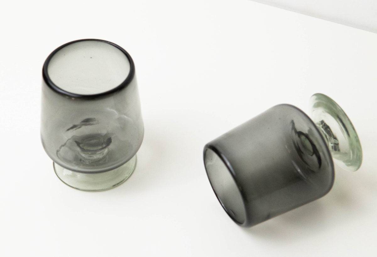 Two short-stemmed wine glasses with dark rim and base, one clear and one smoky gray, displayed against white background.