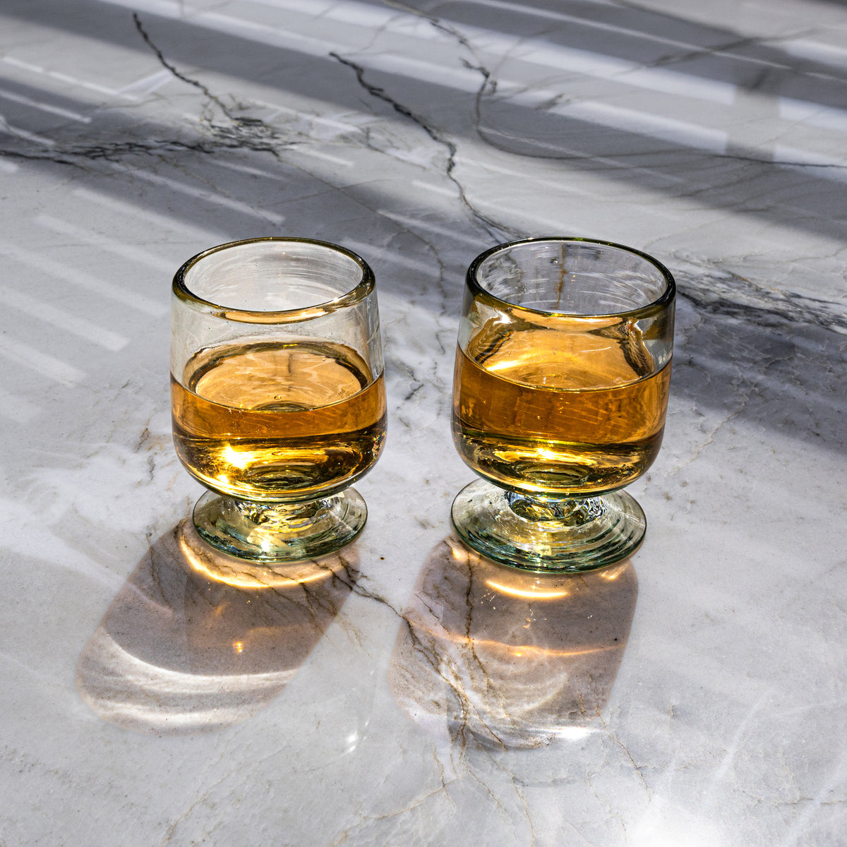 Two short-stemmed wine glasses with gold-tinted rims filled with amber liquid on a marble surface.