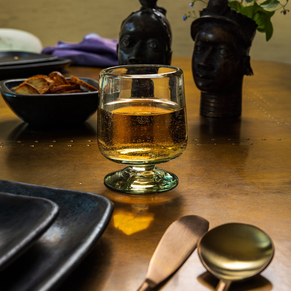 Clear short-stemmed wine glass with amber liquid, displayed on table with snacks and black decorative objects.