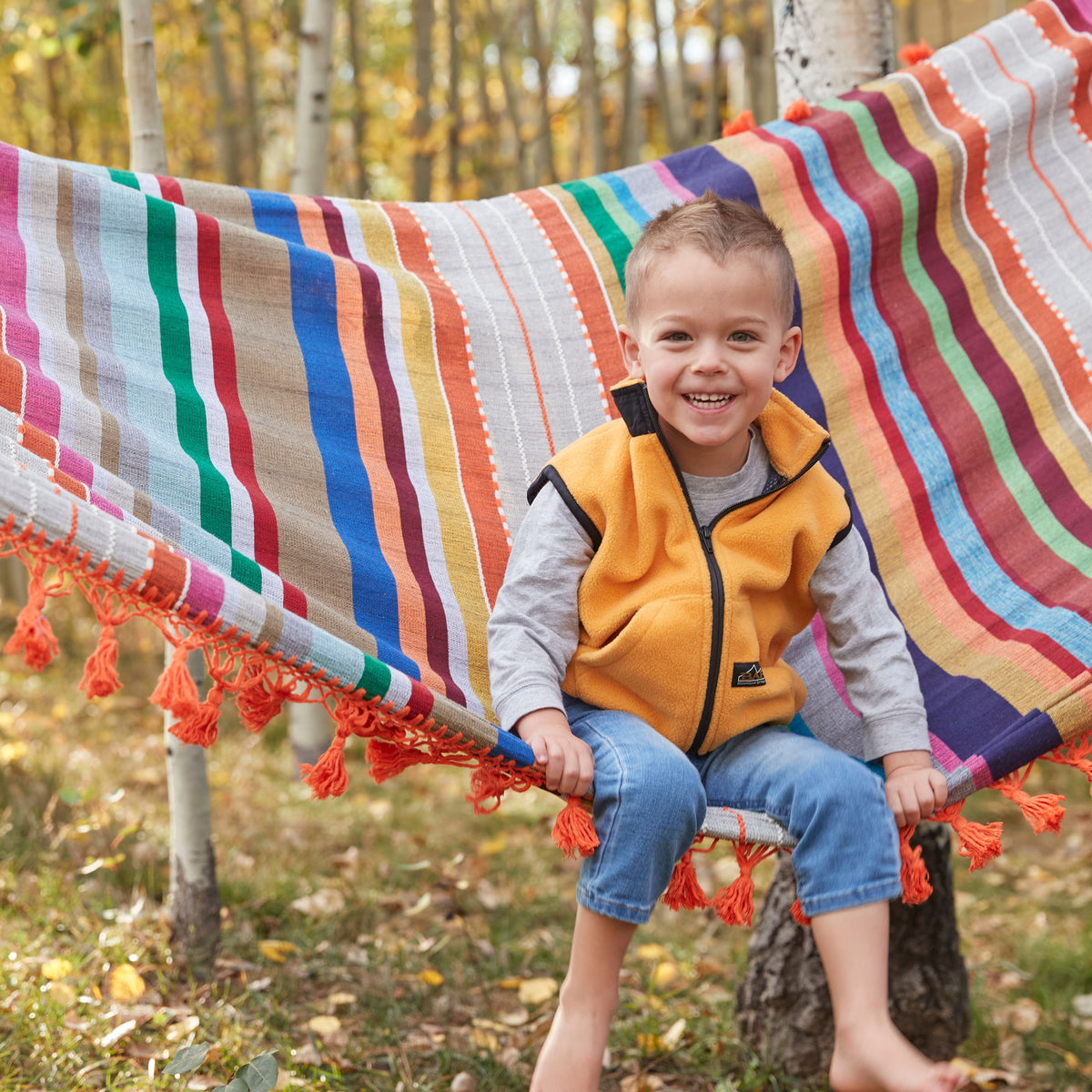 Colorful woven hammock with multicolored horizontal stripes and orange tassel fringe, suspended between trees.