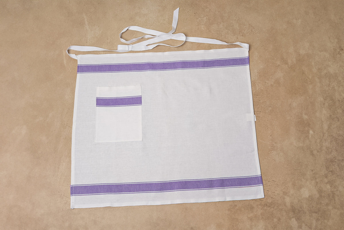 White linen apron with purple trim stripes and a single front pocket, displayed flat with neck ties.
