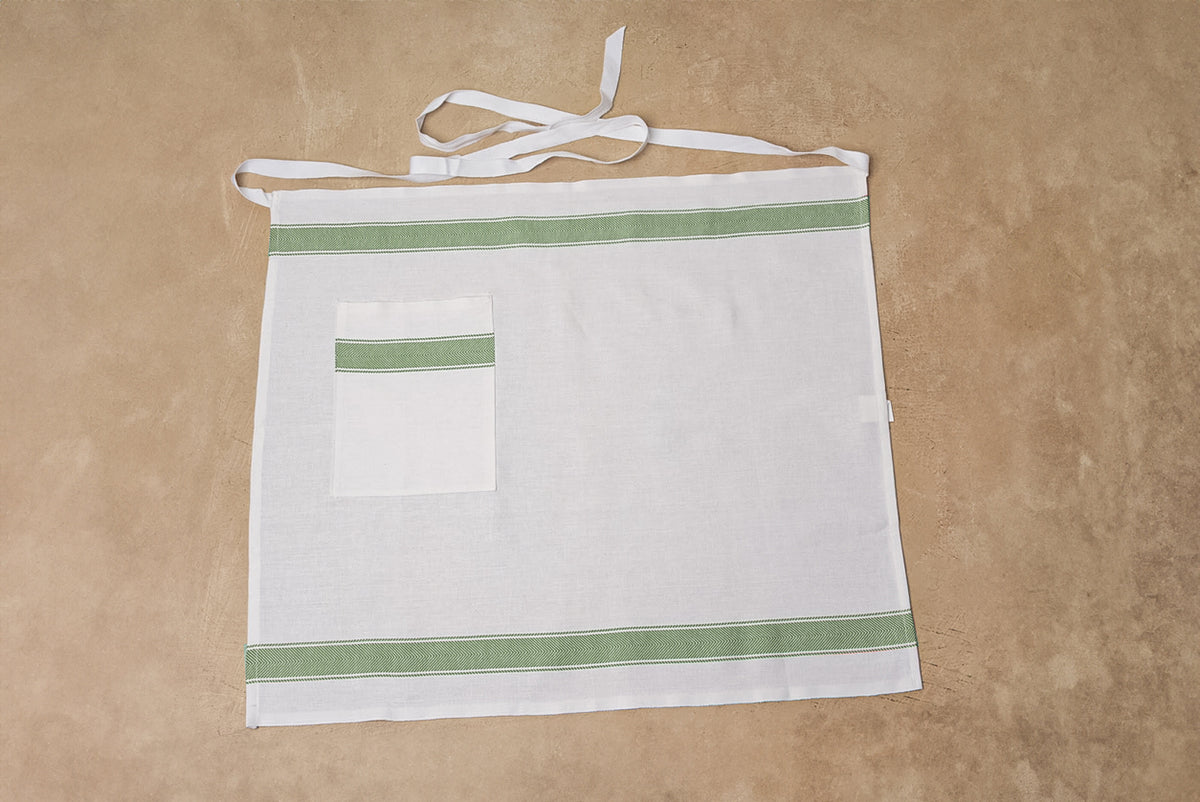 White linen apron with green stripe trim and front pocket, displayed flat with neck and waist ties.