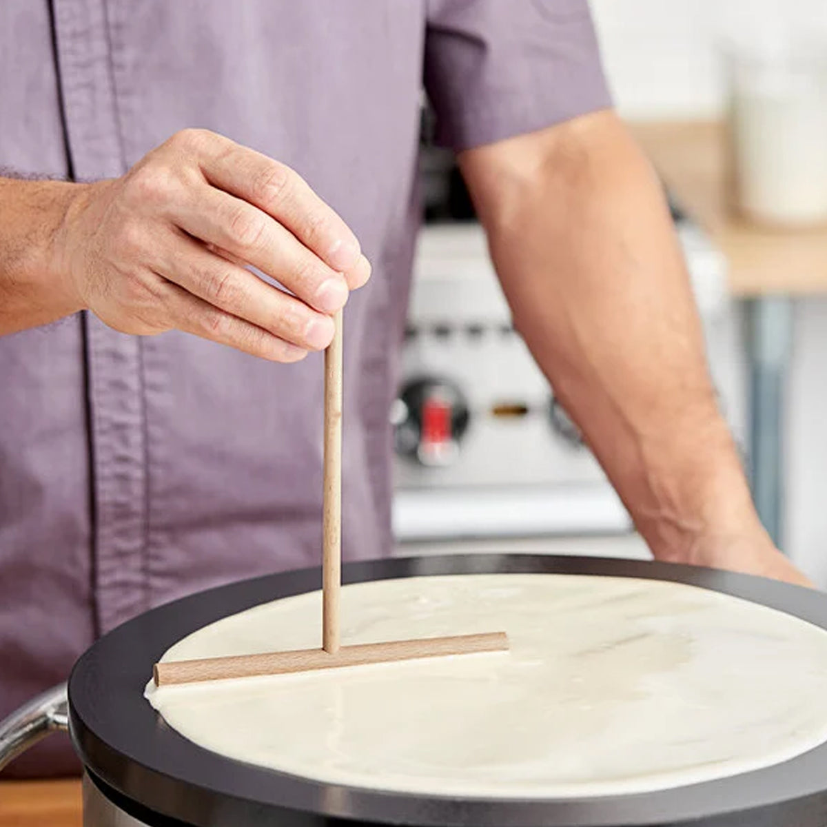 Person holding wooden crepe spreader over black griddle with crepe batter.