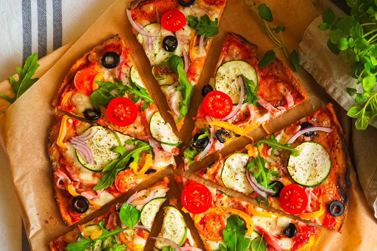 Baked pizza with tomato sauce, cheese, black olives, red tomatoes, zucchini, ham, red onion, and fresh herbs on parchment pap