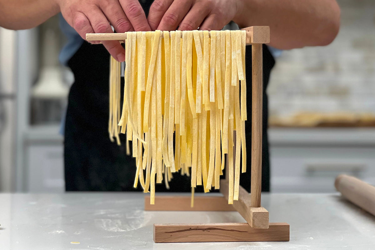 Hands holding wooden drying rack with fresh fettuccine pasta strands draped across it.