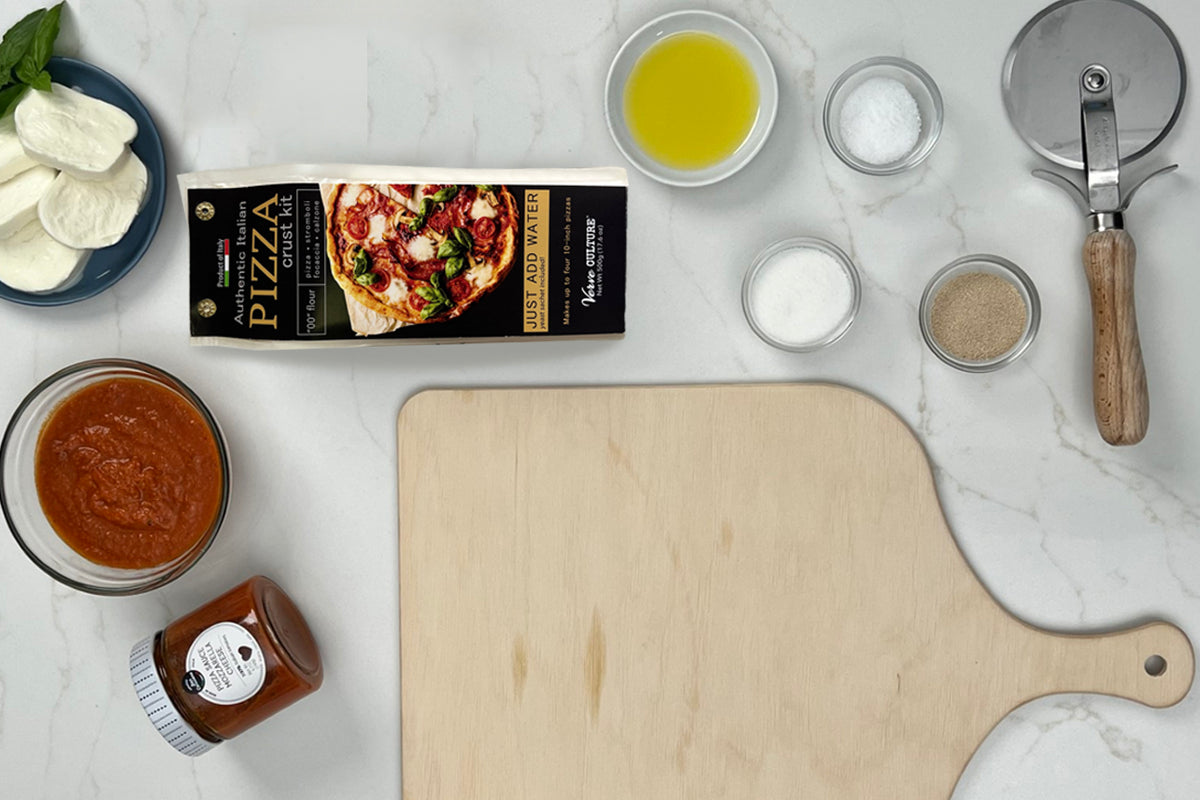 Pizza crust kit with flour box, fresh mozzarella, olive oil, tomato sauce, seasonings, pizza cutter, and wooden serving board