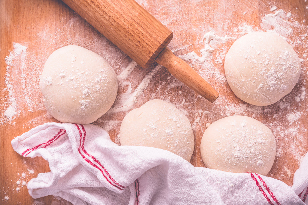 Four balls of pizza dough dusted with flour on a wooden surface with a rolling pin and striped kitchen towel.