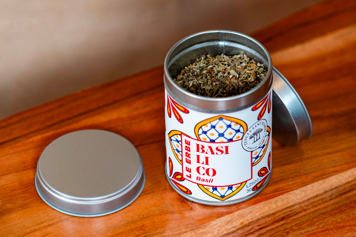 Metal tin container with red and white label filled with dried basil herbs on wooden surface.