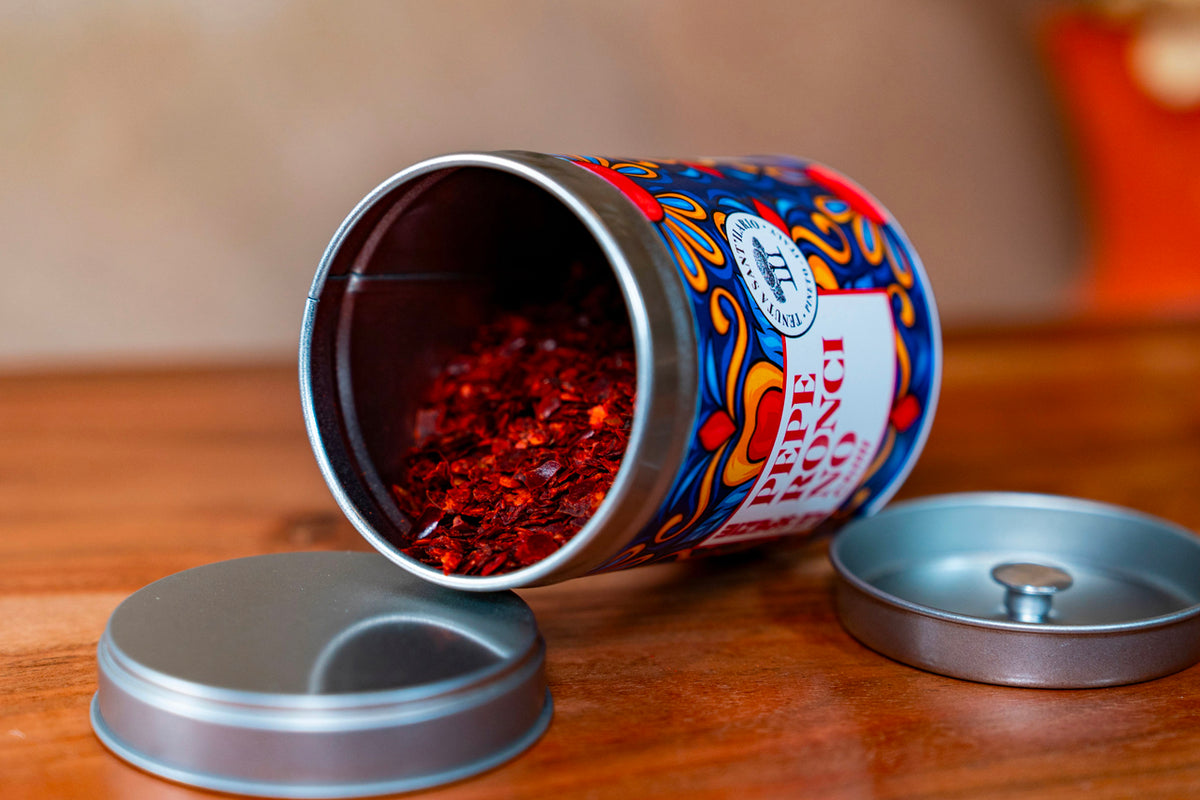 Decorative metal tin with blue and red label tipped over, spilling red dried herbs onto wooden surface with lid beside it.