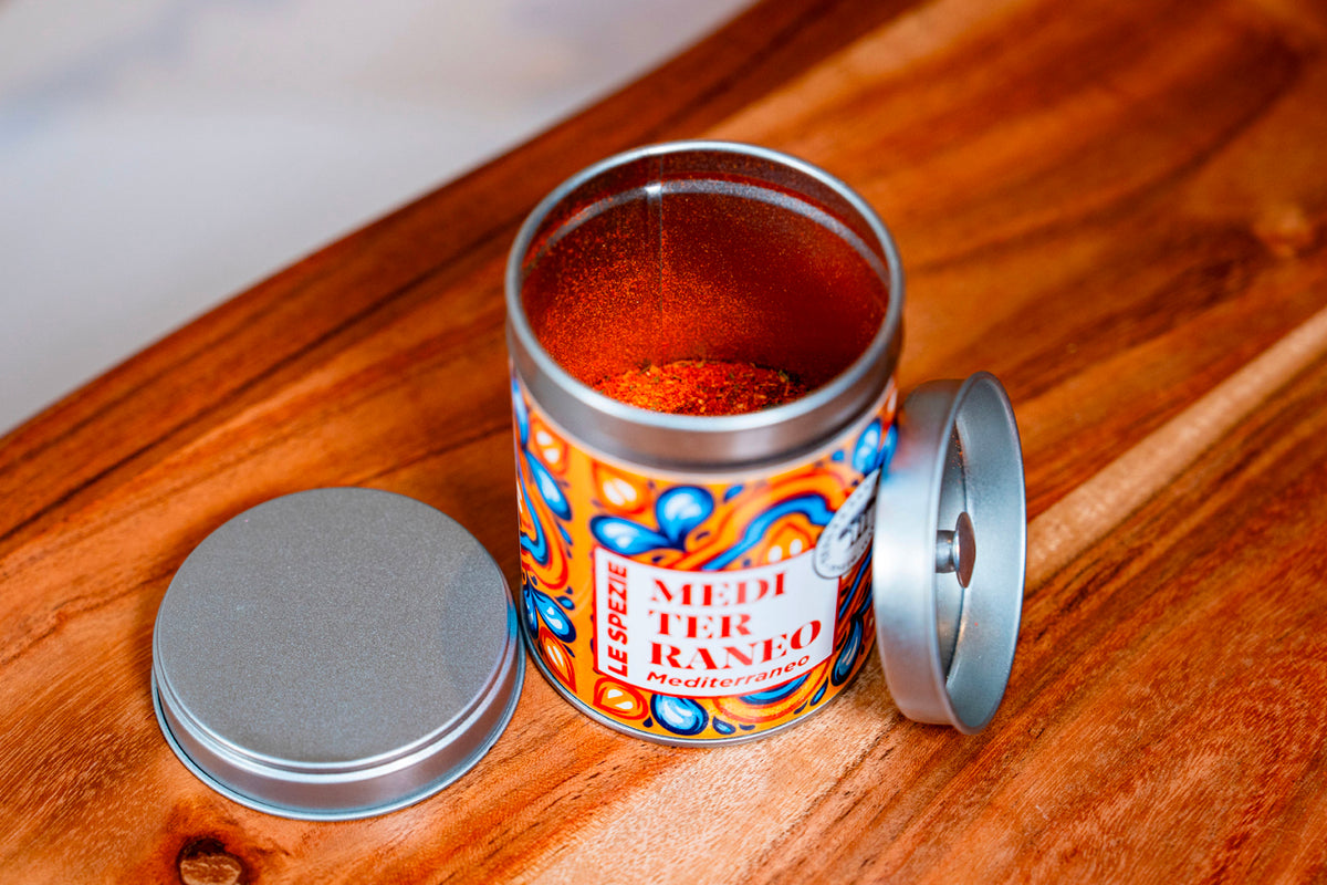 Open spice jar with red dried herb blend and decorative patterned metal container on wooden surface.