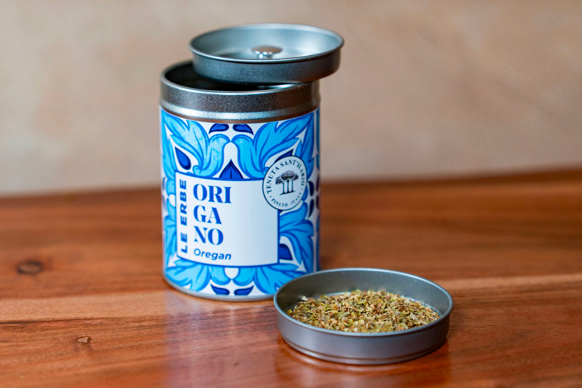 Blue and white decorative tin container labeled "Oregano" with metal lid, beside open tin dish showing dried green herb mixtu