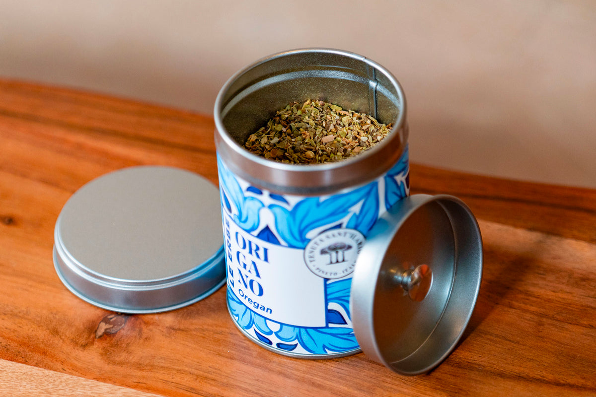Metal tin container with blue and white label filled with dried Italian herb mixture, lid removed and placed beside it on woo