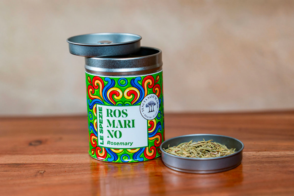 Colorful tin container labeled rosemary with open lid and dried rosemary herbs in small metal dish beside it.