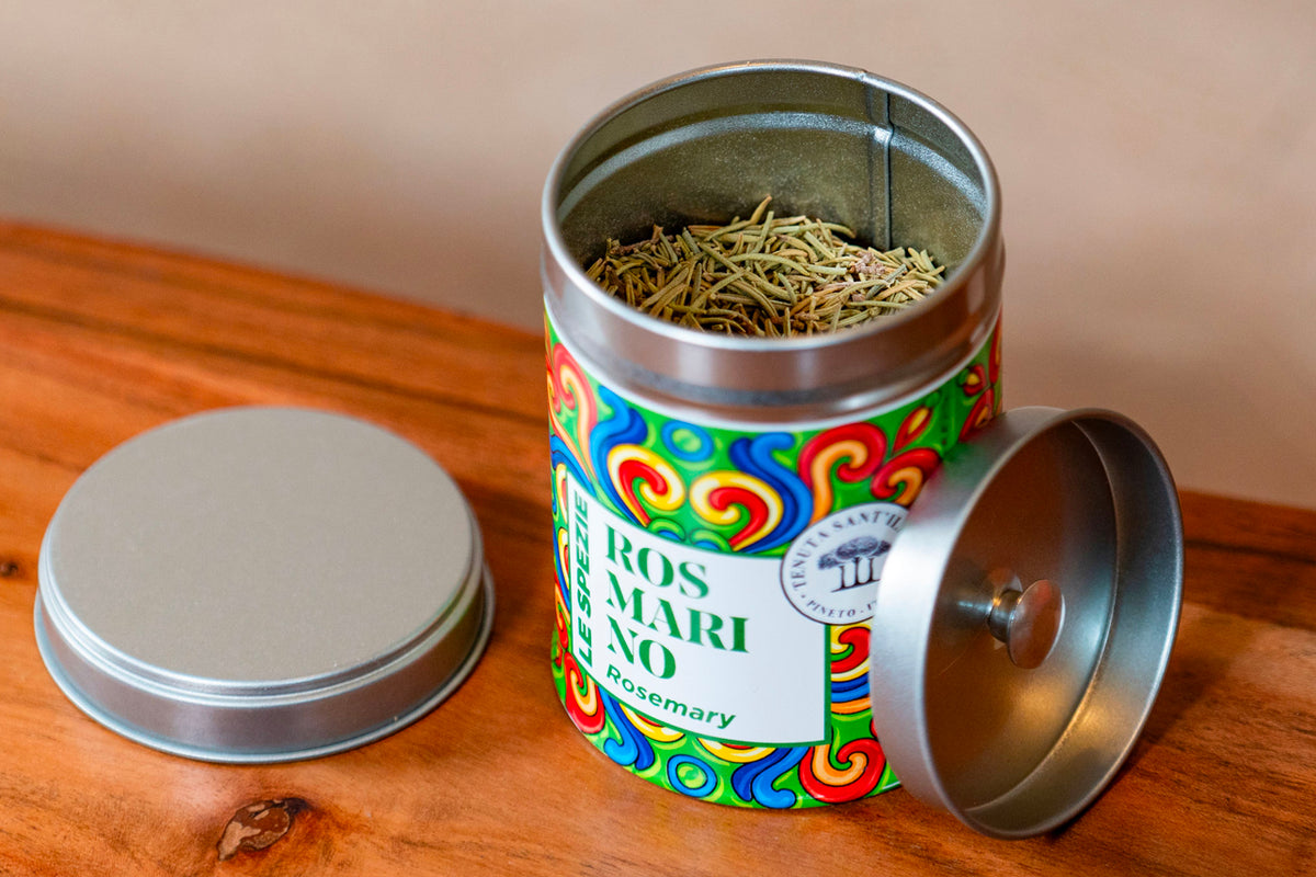 Metal tin container with colorful label containing dried rosemary herbs, shown open with lid removed on wooden surface.