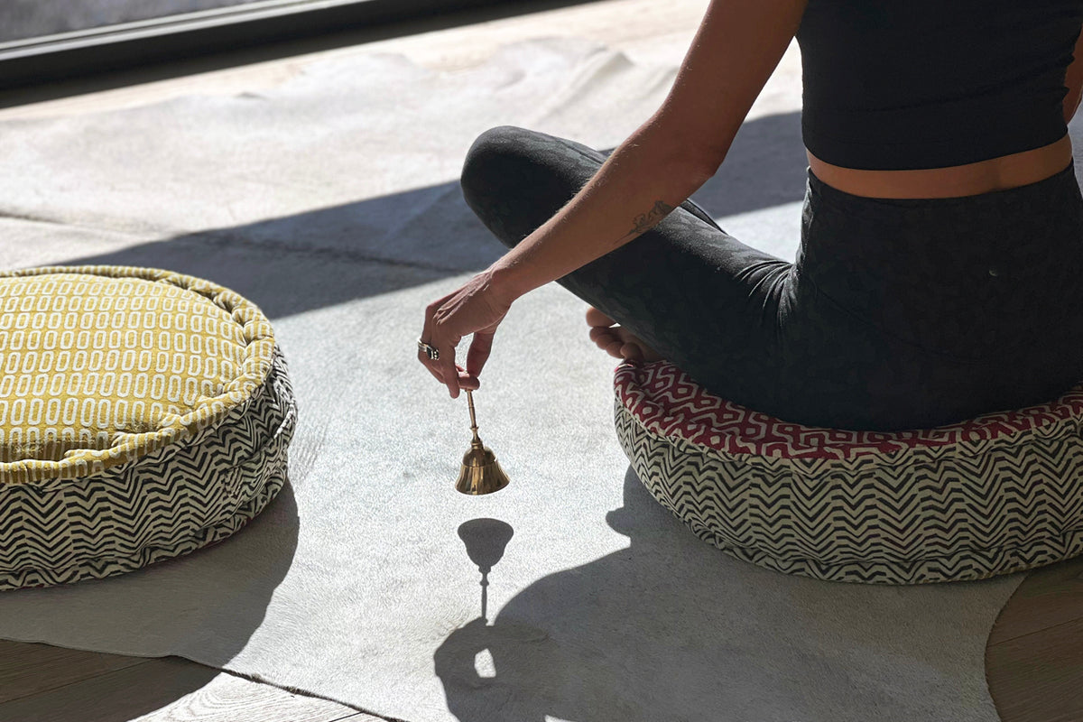 Person holding brass meditation bell with mallet while seated on patterned cushions.