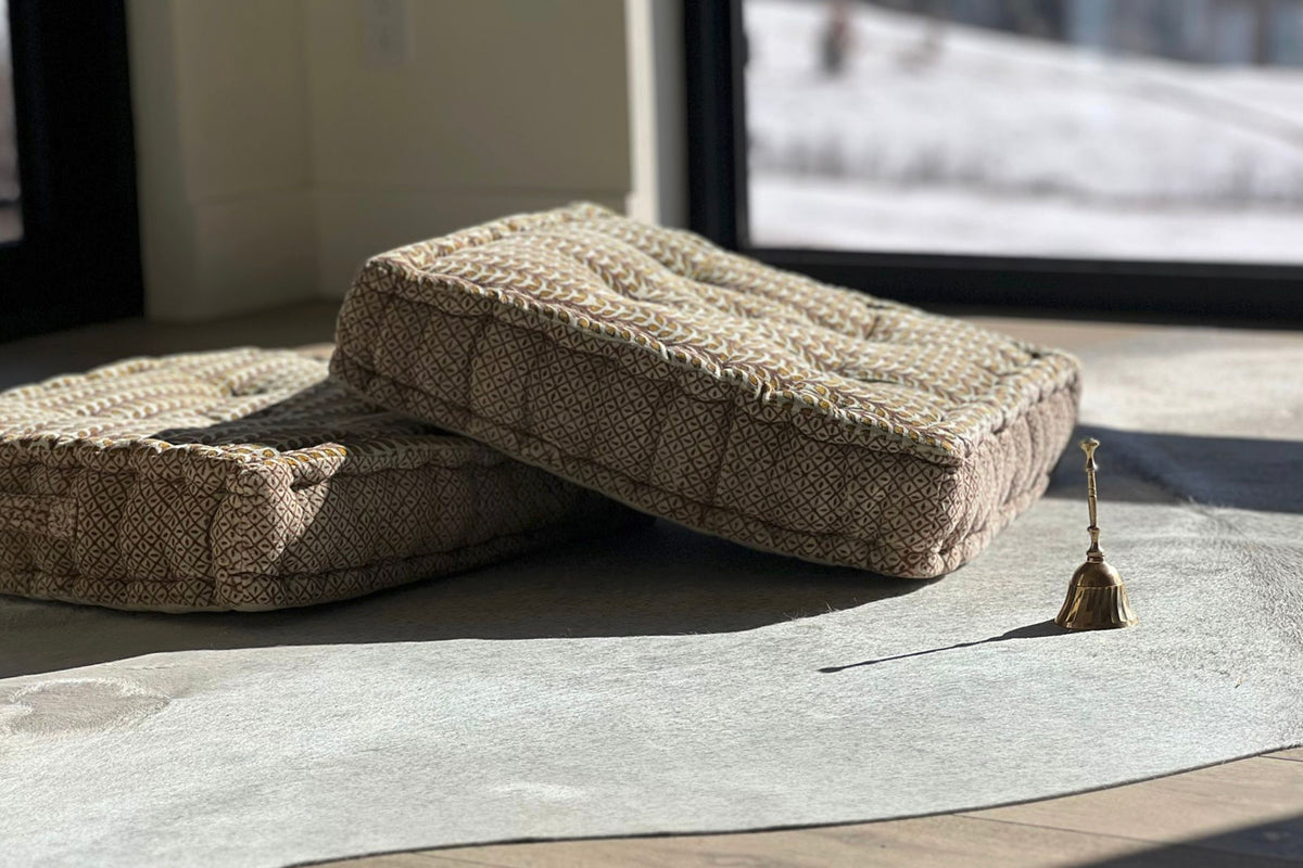 Brass meditation bell with small striker placed on gray mat next to folded natural linen cushions.