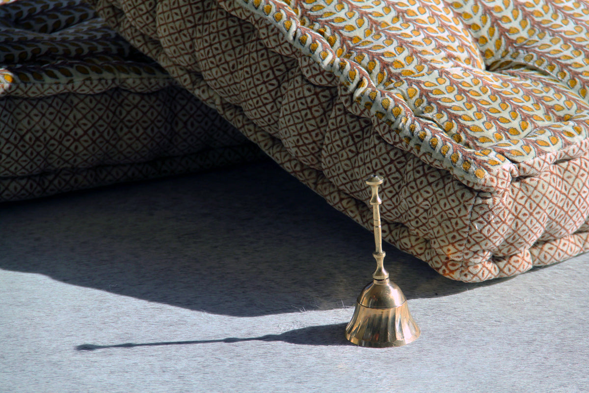 Brass bell with handle on concrete surface next to patterned fabric cushions.