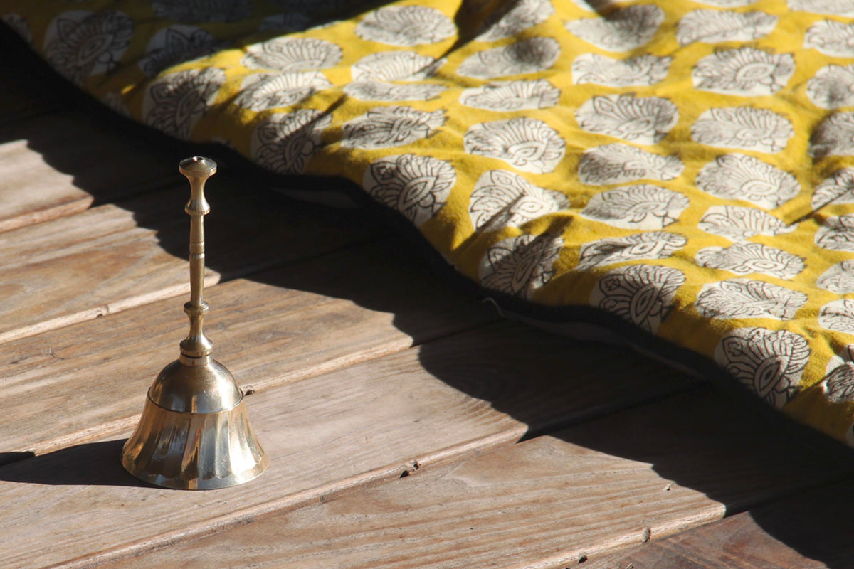 Brass bell with handle on wooden surface next to yellow patterned cushion.