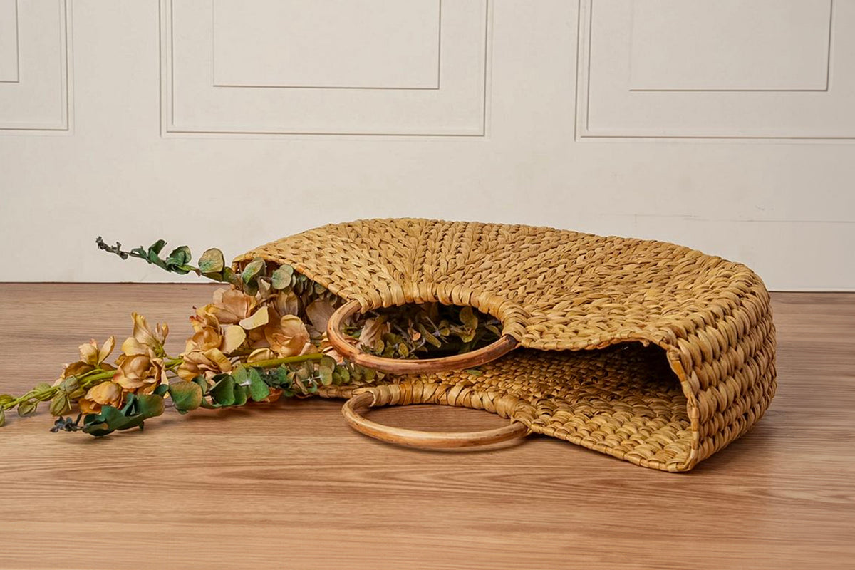 Woven straw tote bag with circular wooden handles displayed on wood flooring next to dried flowers.