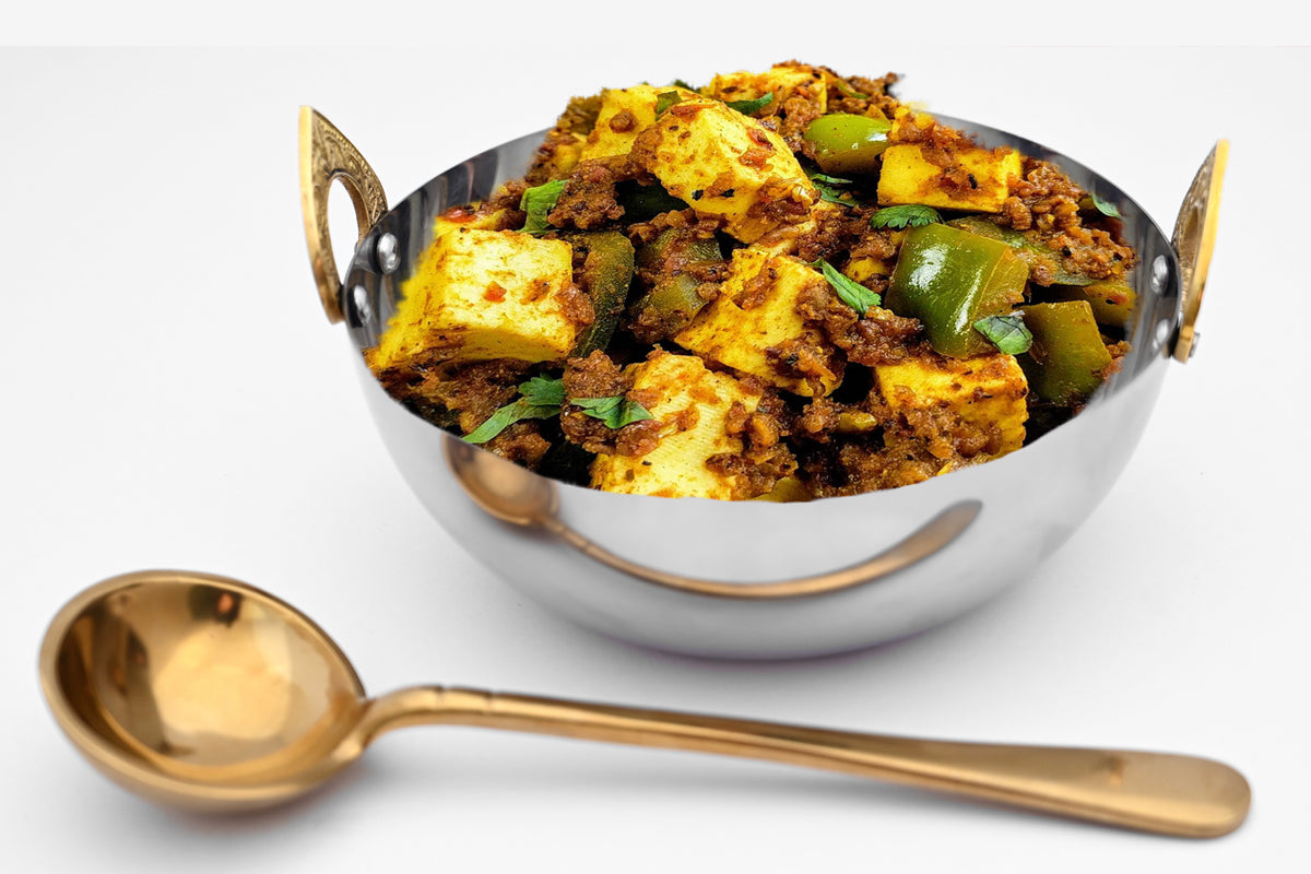 Stainless steel handi bowl with brass handles filled with spiced curry and vegetables, shown with matching brass serving spoo