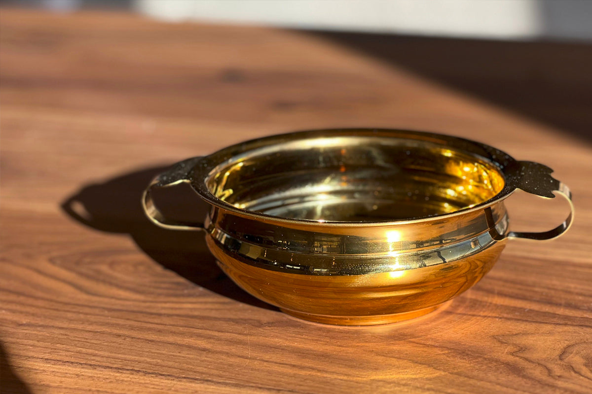 Brass-colored stainless steel handi bowl with two handles on a wooden surface.