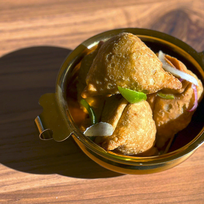 Brass-colored stainless steel handi bowl containing fried dough balls with spices and herbs on wooden surface.