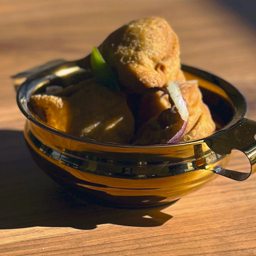 Brass-colored stainless steel handi bowl with handles containing fried food, onion slice, and green garnish.