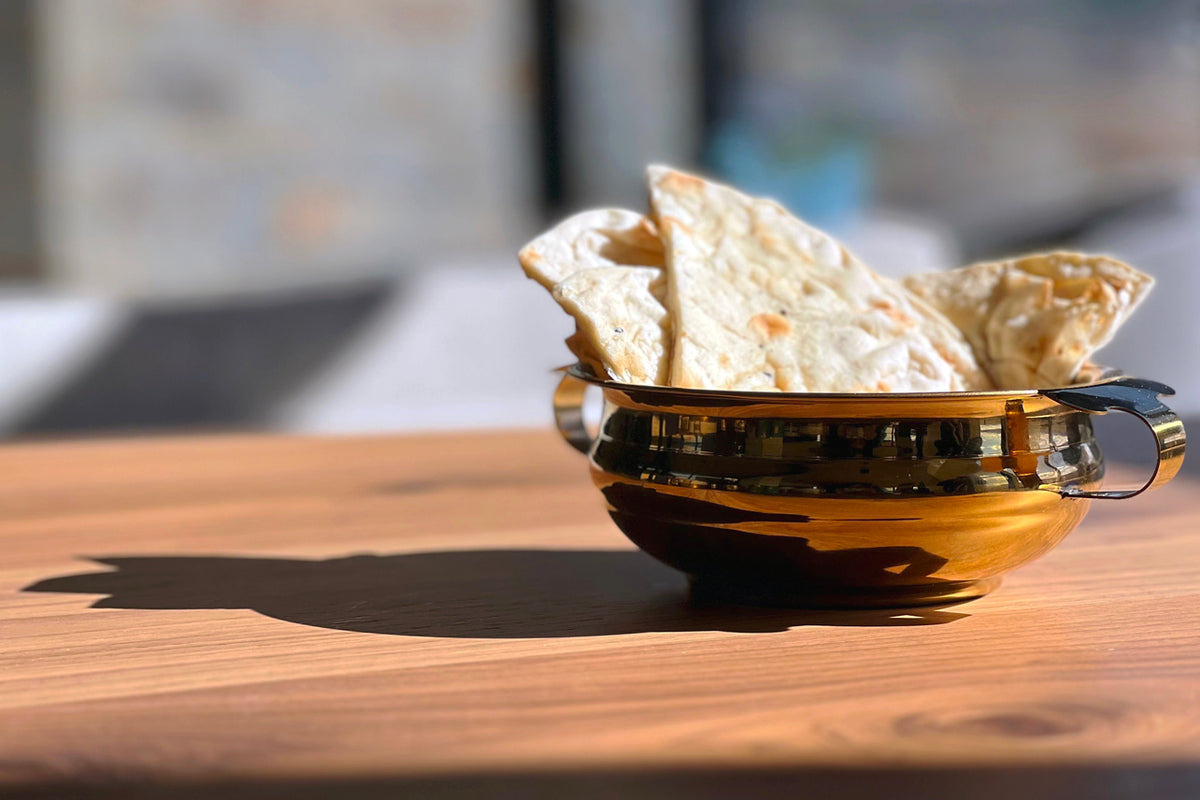 Brass-colored stainless steel handi bowl with two handles holding flatbread on wooden surface.