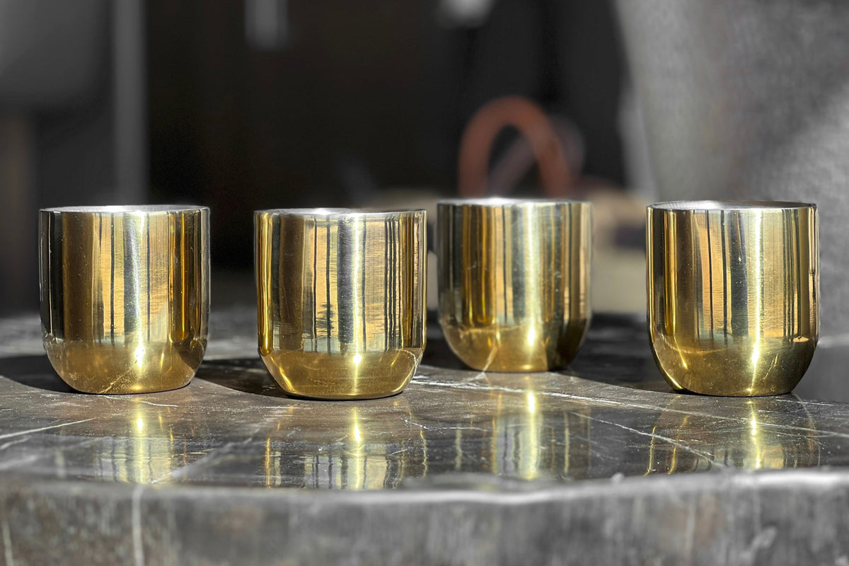 Four brass-colored double-walled cups with cylindrical shape arranged in a row on a marble surface.