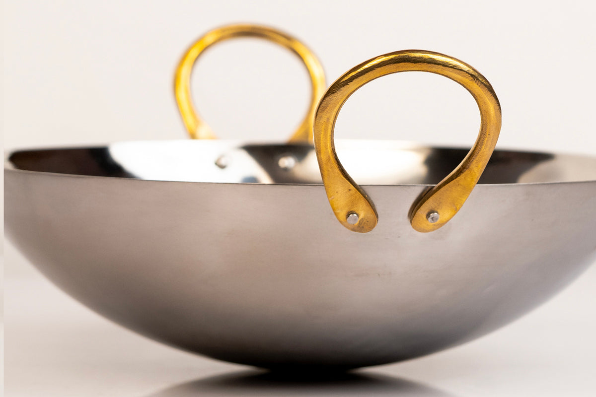 Stainless steel kadai wok with two curved brass handles on opposite sides.