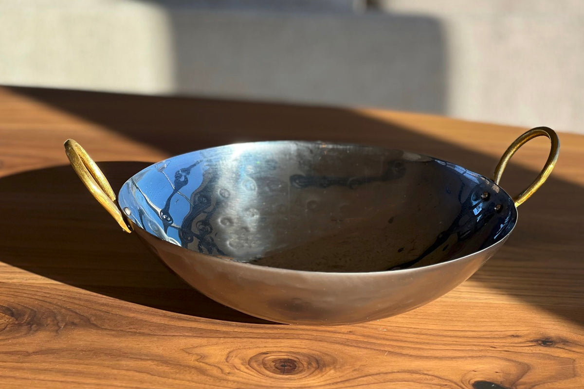 Stainless steel wok with curved sides and brass loop handles on each side, sitting on a wooden surface.