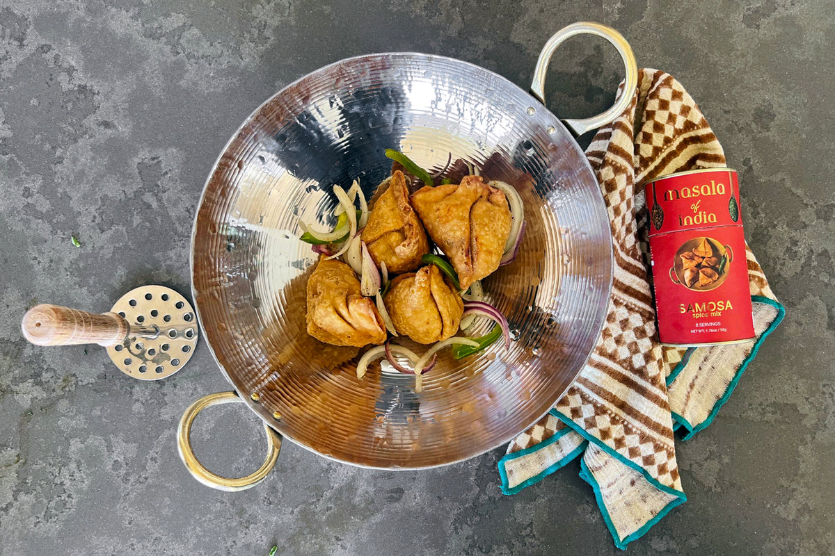 Stainless steel kadai wok with brass handles containing cooked vegetables and onions, displayed with a wooden spoon and kitch