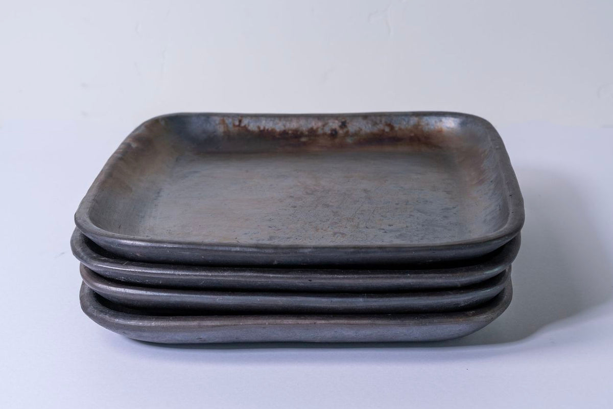 Stack of four dark gray handmade ceramic dinner plates with rounded square shape and shallow wells.